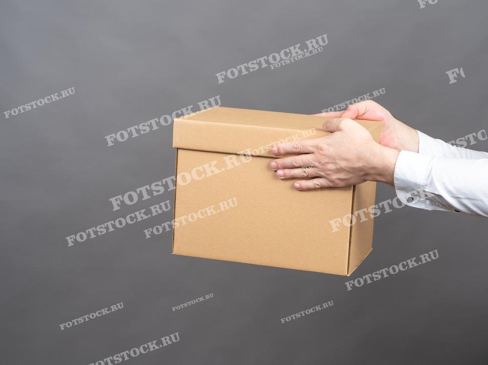 На фотографии изображены руки, держащие картонную коробку на фоне серой стены. Снимок подчеркивает процесс перемещения и хранения личных вещей на складе.
